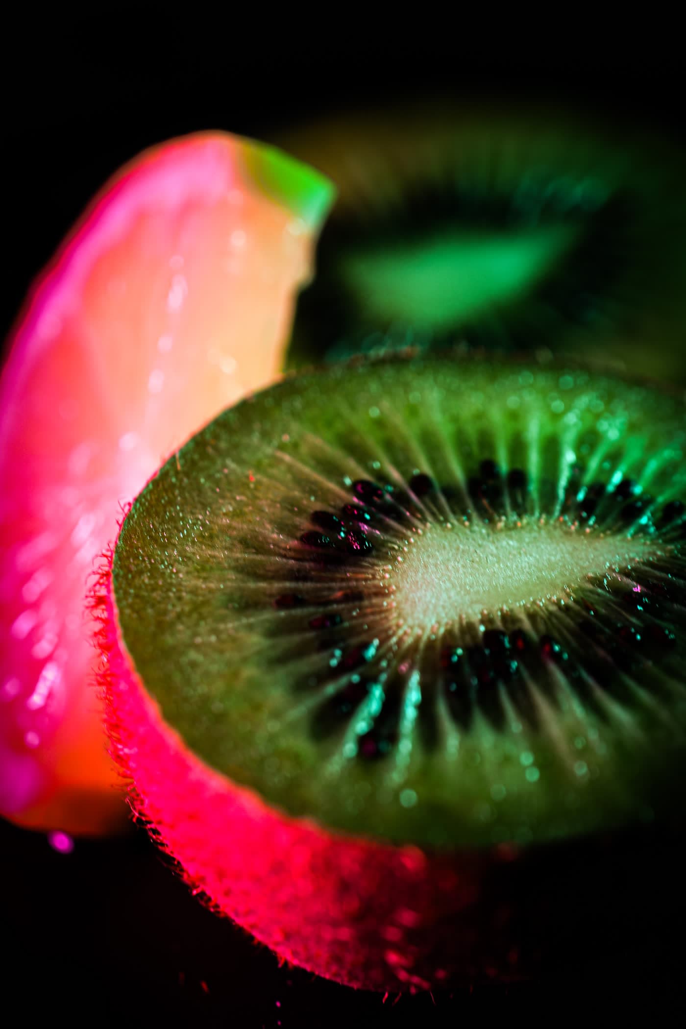 A slice of kiwi fruit lit by pink and green light against a black background.