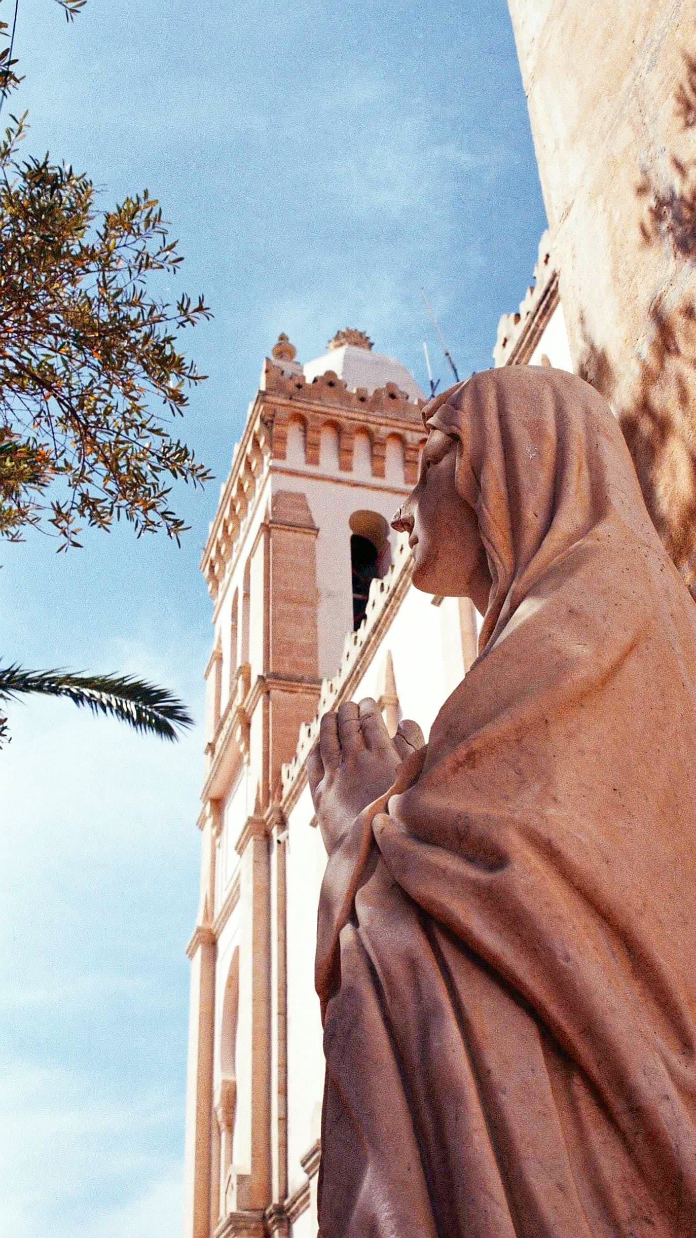 A stone statue in a hooded robe with hands together, beside a pale church tower under a blue sky.