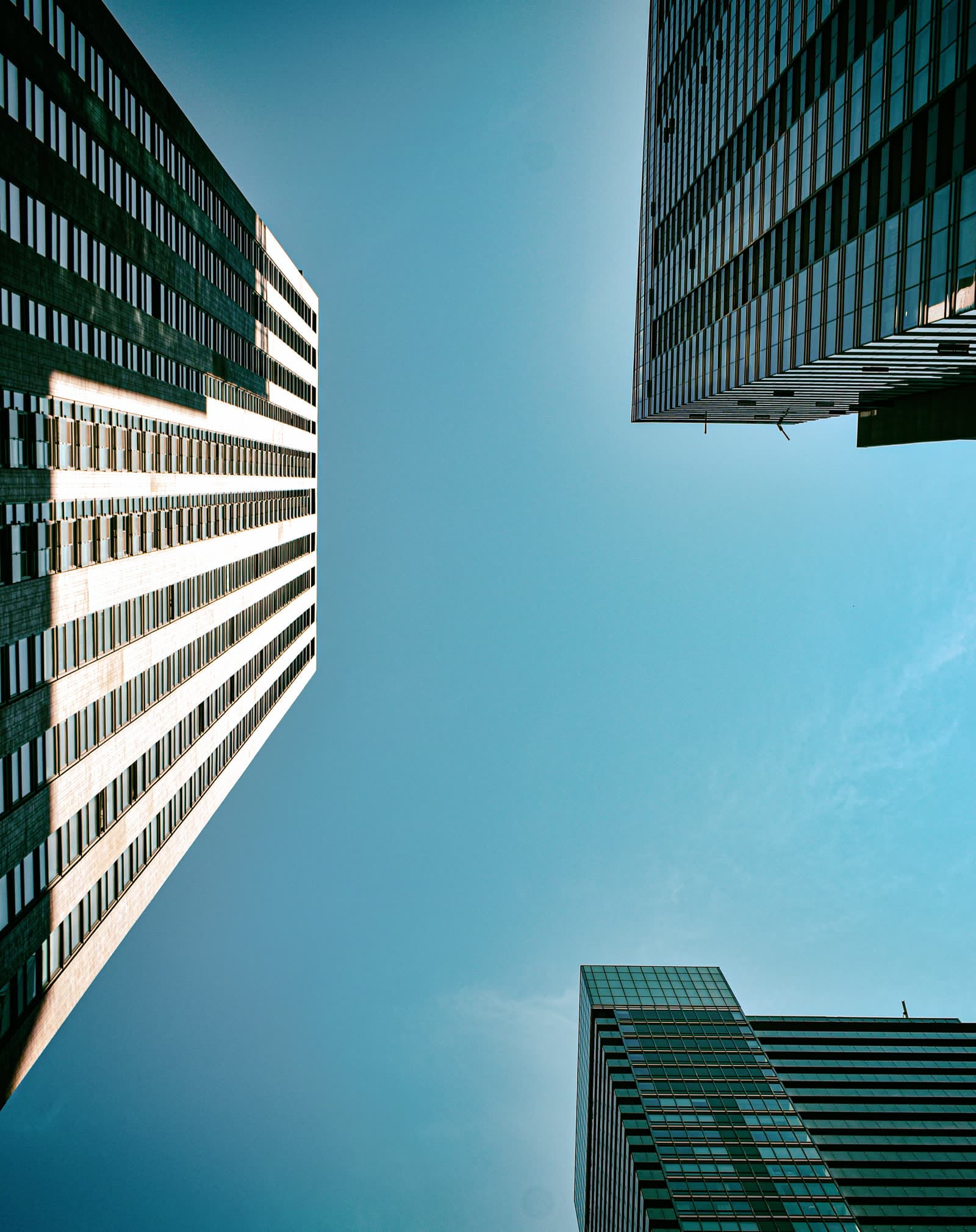 Three glass office towers photographed from street level, converging into a clear teal sky.