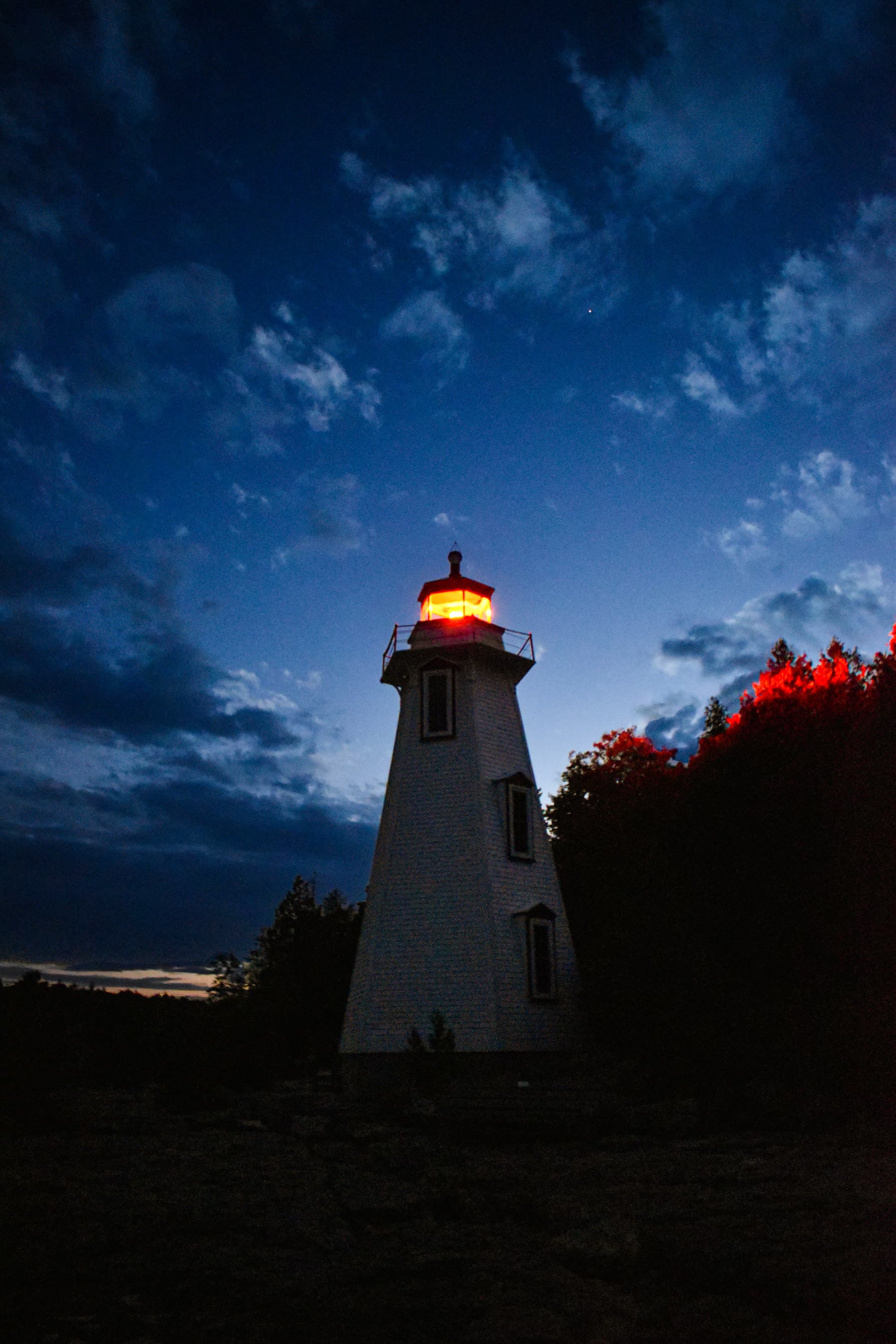 A small white lighthouse at blue hour, its lamp burning orange against a deep blue sky full of cloud.