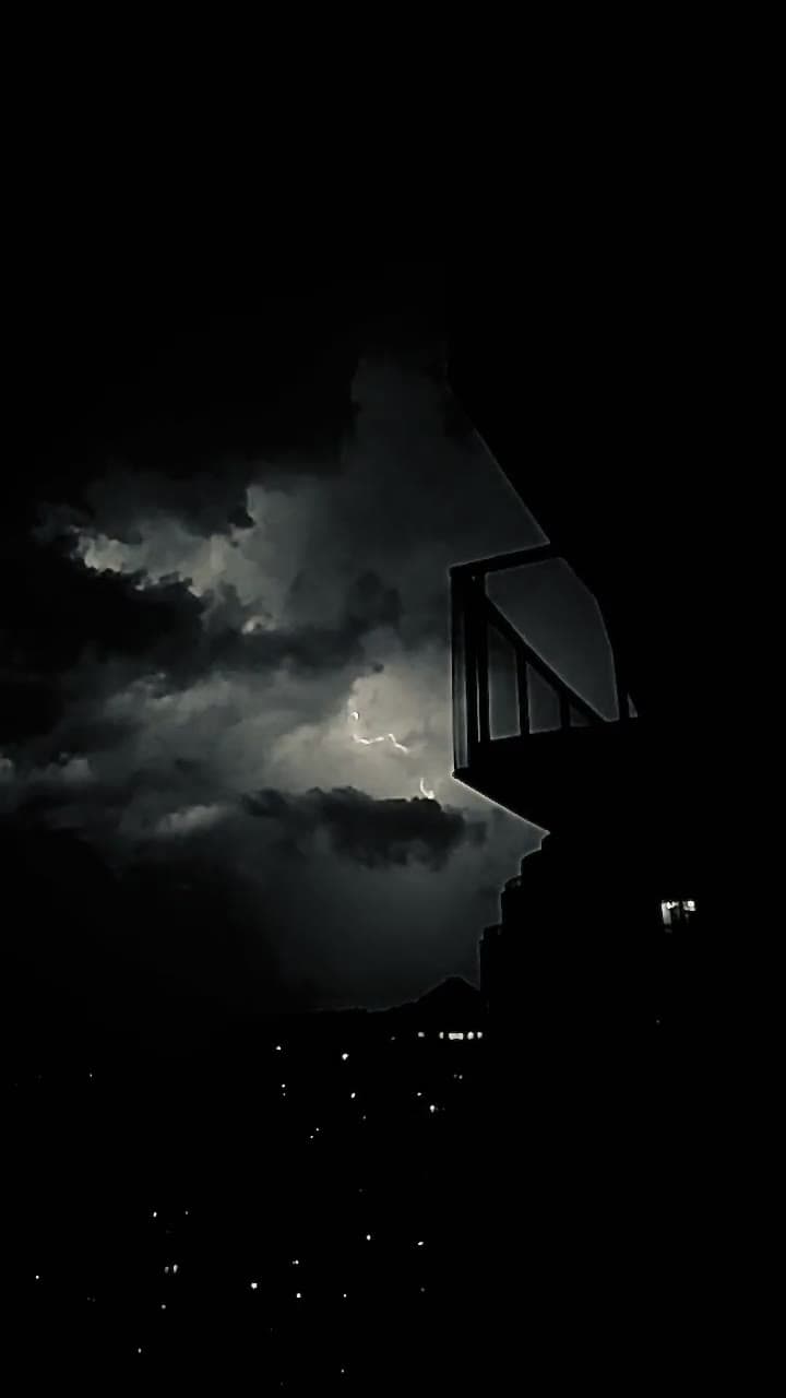 A dark city skyline at night with lightning showing inside cloud beside a balcony railing.